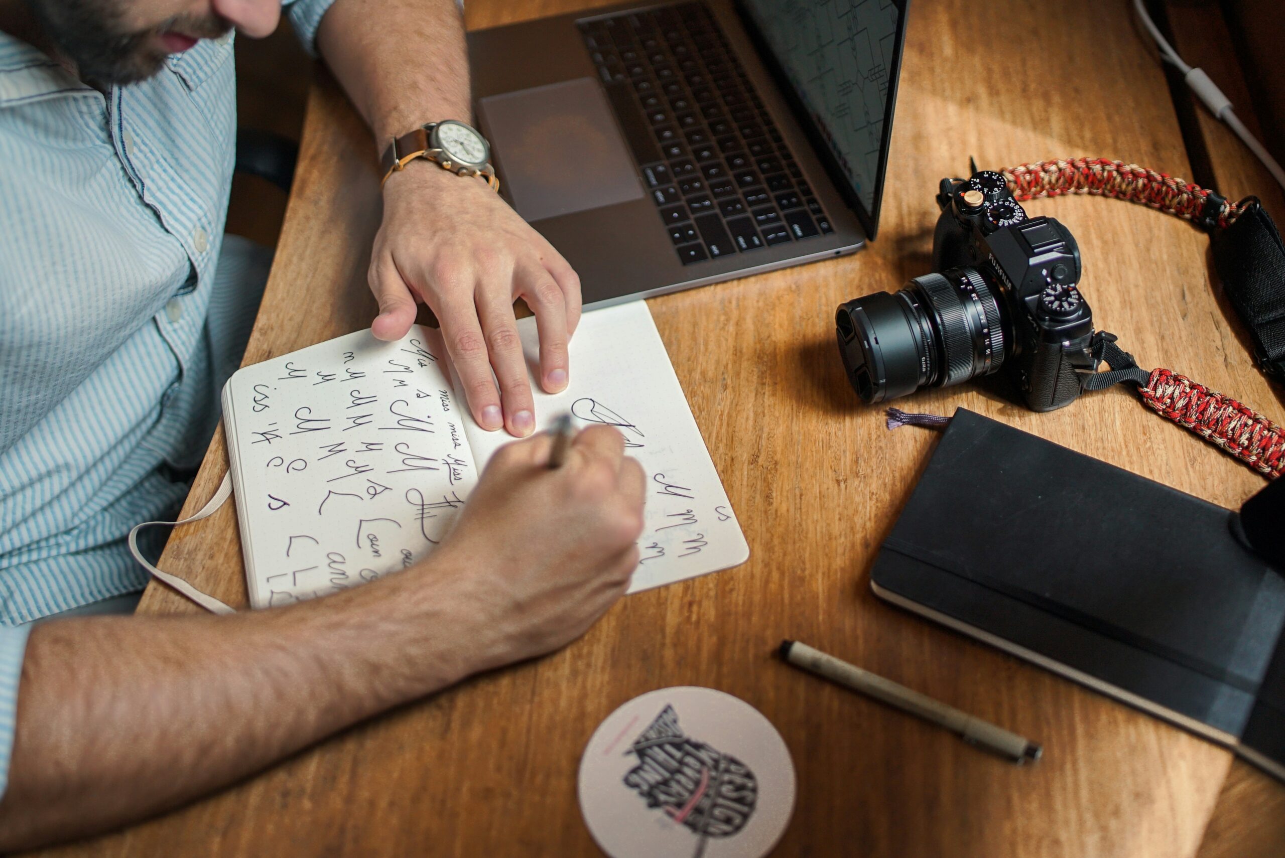 Person writing in notebook next to camera and laptop.